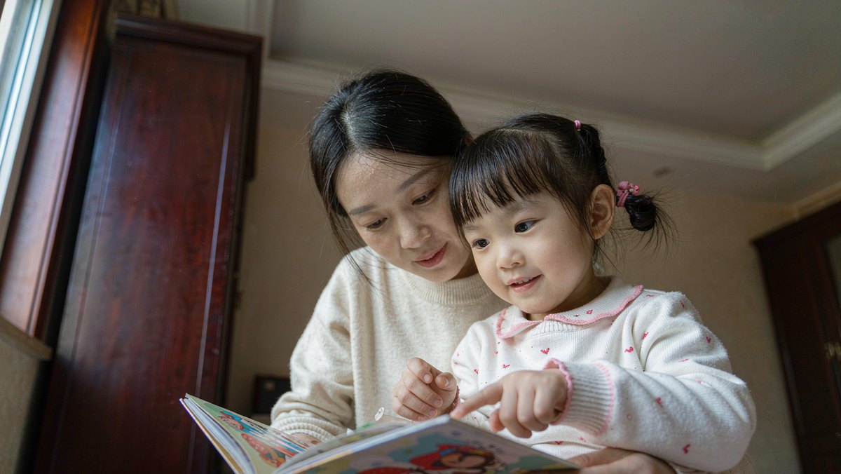 妈妈和女儿并排翻看绘本，孩子指着图画