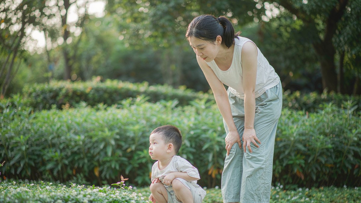 小男孩蹲在花园里看一只停在花上的蝴蝶，妈妈在旁边看