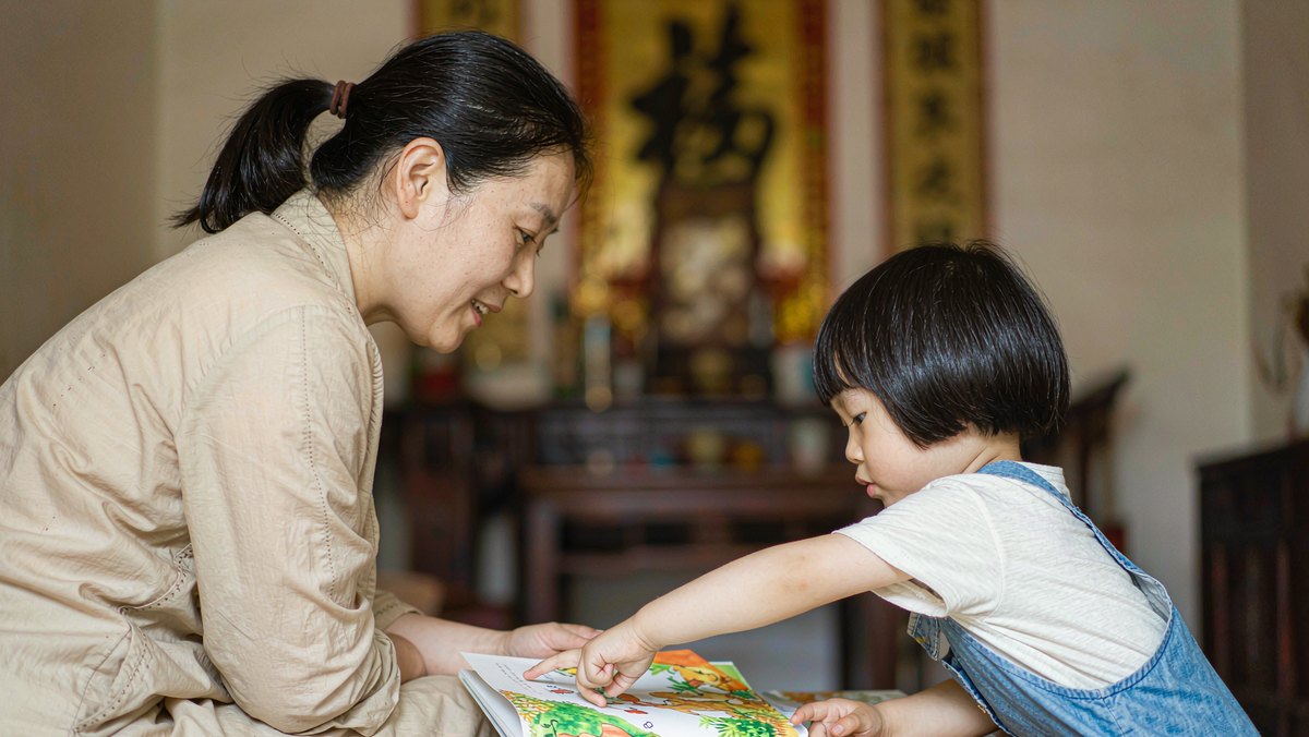 妈妈和孩子在绘本前，孩子伸手要翻页，妈妈笑着等他