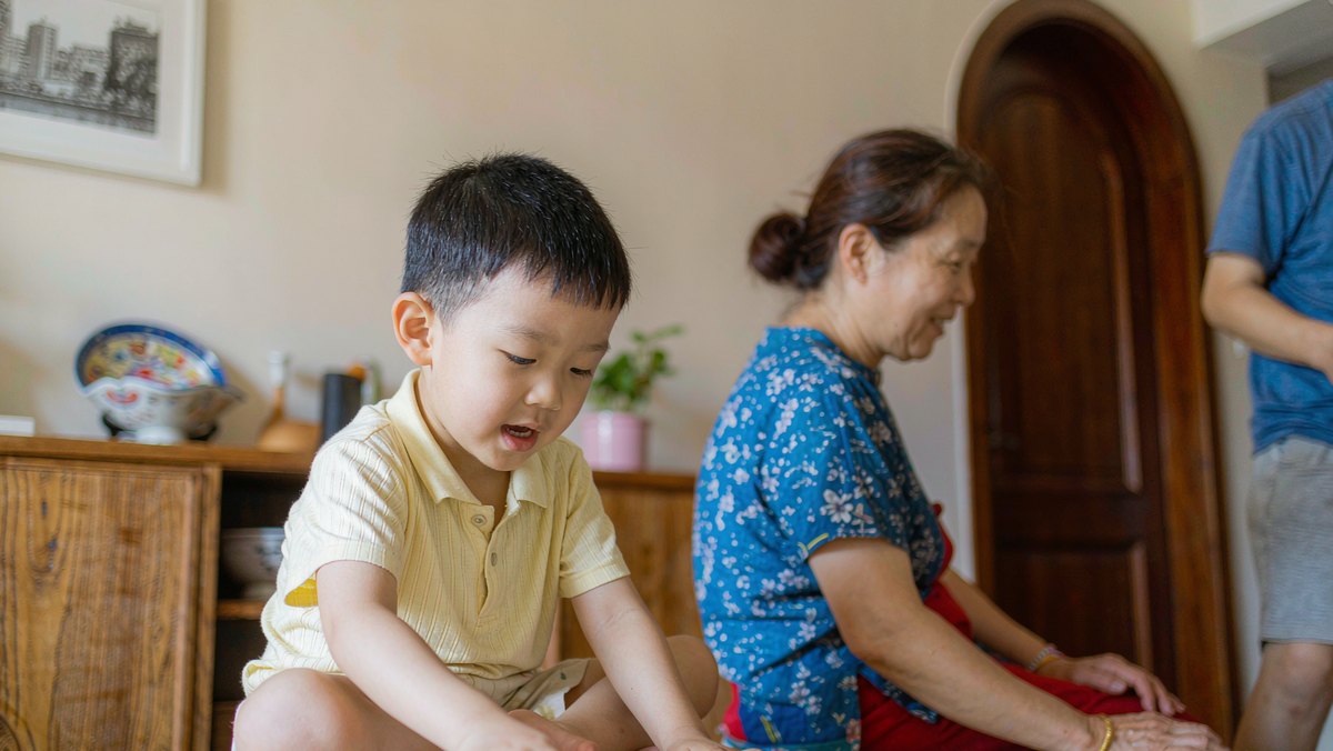 孩子坐在地垫上对着绘本自言自语，妈妈在旁边温柔看着